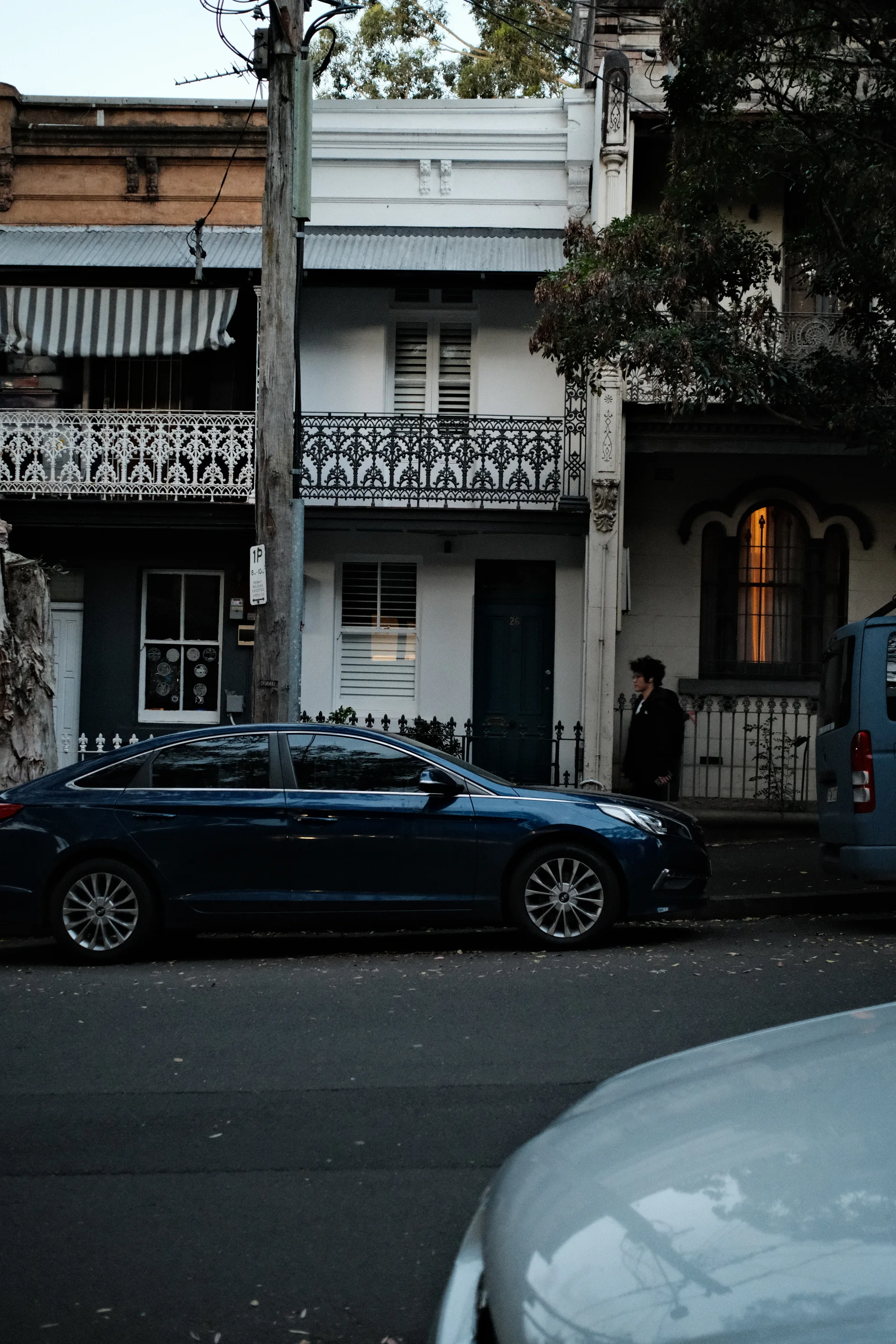 A dark blue car parked outside a narrow white terrace house at dusk.